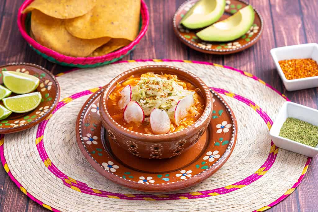 Cibo messicano tradizionale.  Zuppa di pozole rossa con pollo accompagnata da avocado, lattuga, cipolla, ravanello, limone, peperoncino e tortillas di mais croccanti conosciute anche come tostadas su sfondo di legno.