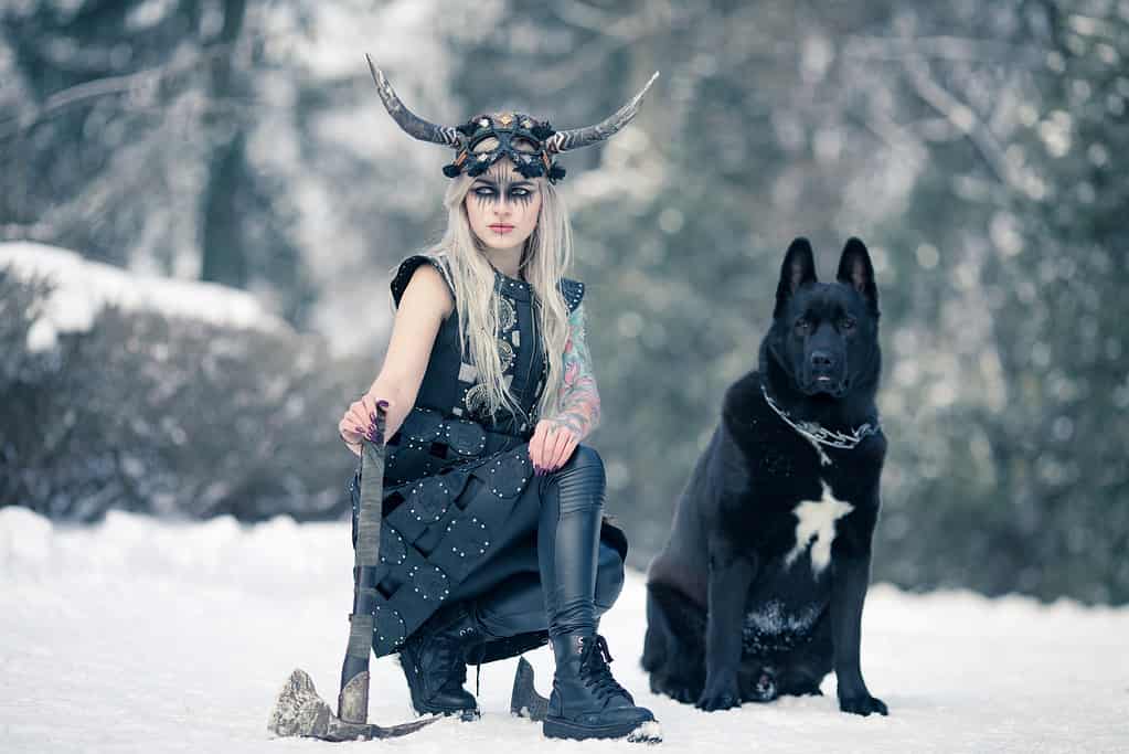 Donna guerriera nell'immagine del vichingo con ascia ed elmo cornuto accanto al grosso cane nero nella foresta invernale.