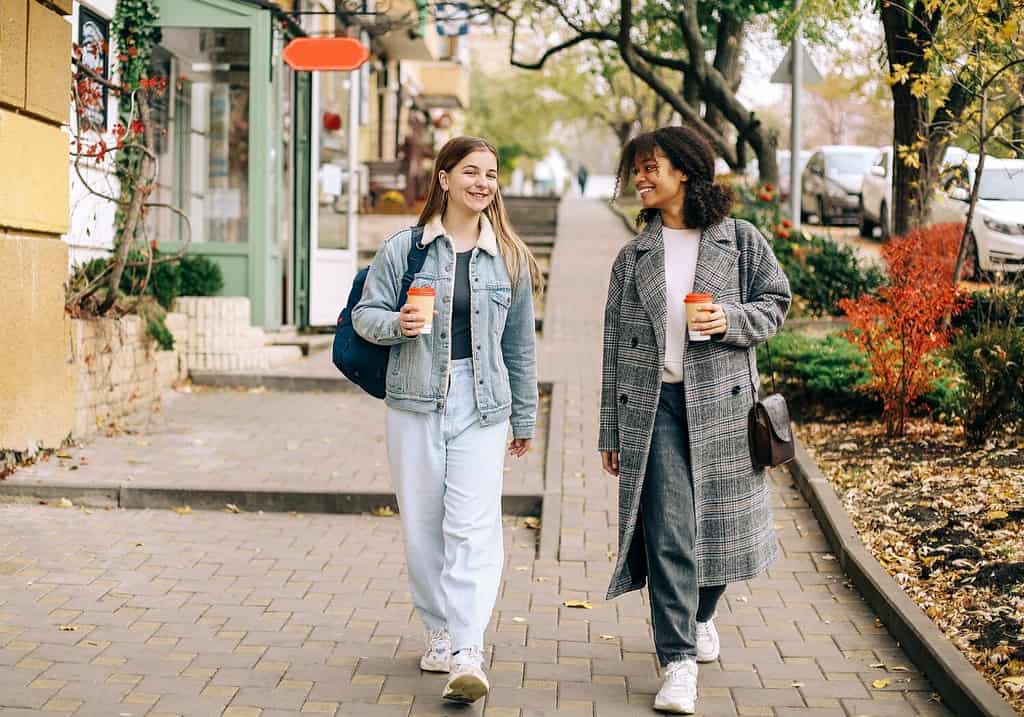 Due amiche multirazziali felici e spensierate che camminano per le strade della città con caffè da asporto, parlando e condividendo storie di vita, migliori amiche multietniche che si godono una passeggiata autunnale all'aperto.  L'amicizia delle donne