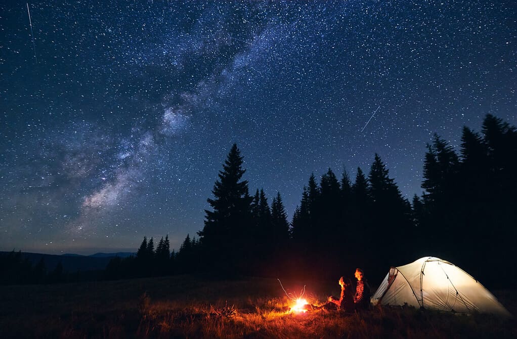Giovane coppia di escursionisti seduti vicino al luminoso falò acceso e alla tenda turistica illuminata, godendosi la notte di campeggio insieme sotto il cielo scuro pieno di stelle lucenti e luminosa Via Lattea, calda serata estiva.