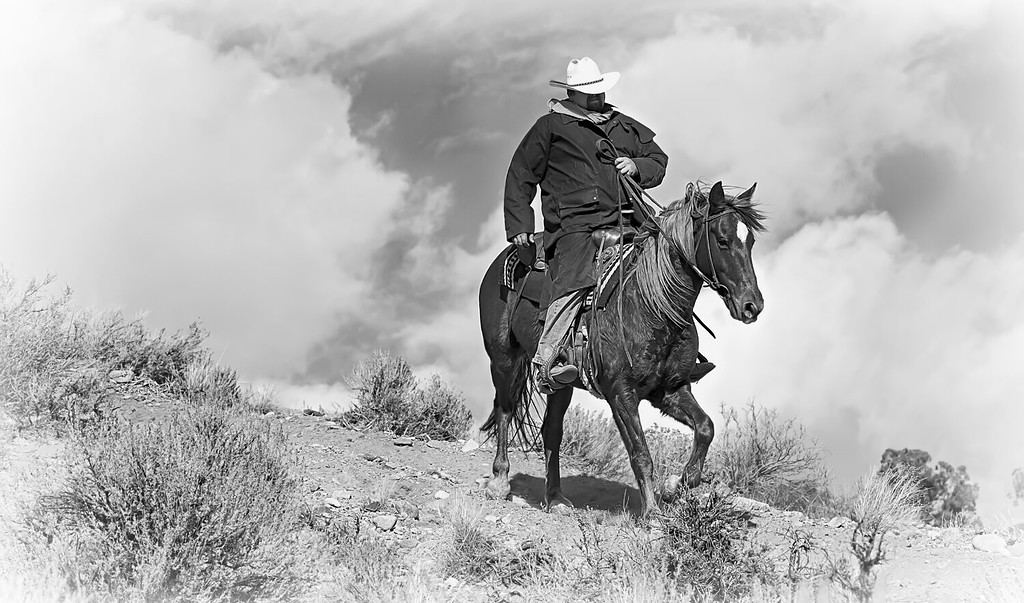 Pony Express Ridge Rider 1 in bianco e nero.  Un cowboy solitario con un cappotto nero cavalca il suo cavallo lungo un crinale montuoso con cieli drammatici dietro di lui.