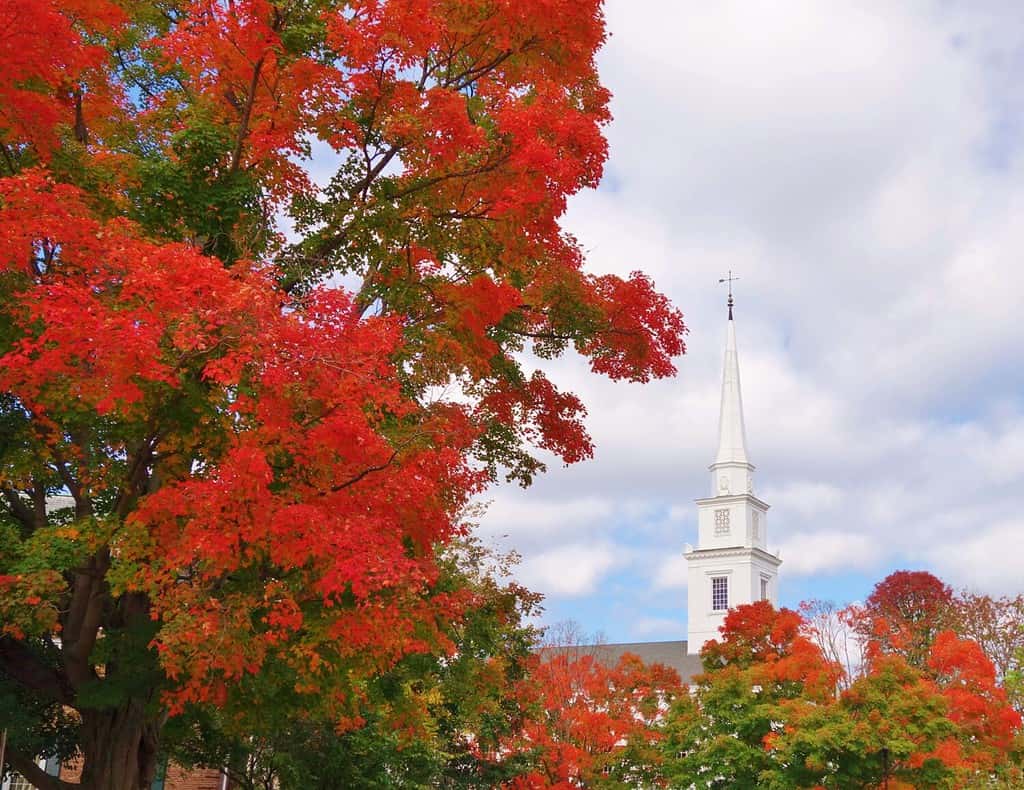 Un grande bellissimo albero rosso brillante dai colori autunnali riempie la metà sinistra del fotogramma con il campanile di una chiesa sotto un cielo azzurro con nuvole bianche gonfie sulla destra ad Hanover, nel New Hampshire, in una bella giornata autunnale