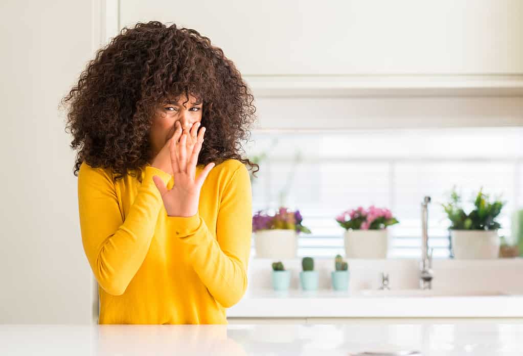Donna afroamericana che indossa un maglione giallo in cucina che sente l'odore di qualcosa di puzzolente e disgustoso, intollerabile, trattenendo il respiro con le dita sul naso.  Concetto di cattivi odori.