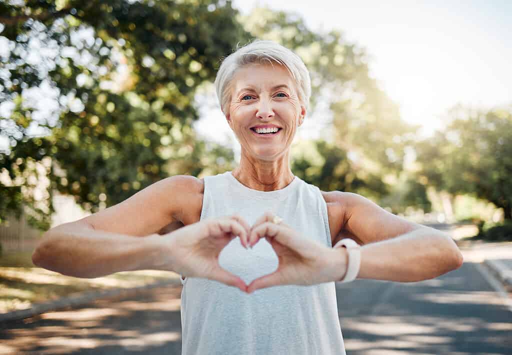 Mani fitness, felici e cardiache di un'anziana nella natura dopo aver corso per salute, benessere e allenamento.  Sorridi, motivazione e pace con la signora anziana e segno di amore, fede e formazione nella natura