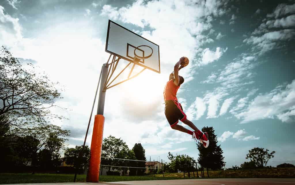 Giocatore di basket di strada che fa una potente schiacciata sul campo - allenamento atletico maschile all'aperto al tramonto - concetto di sport e competizione