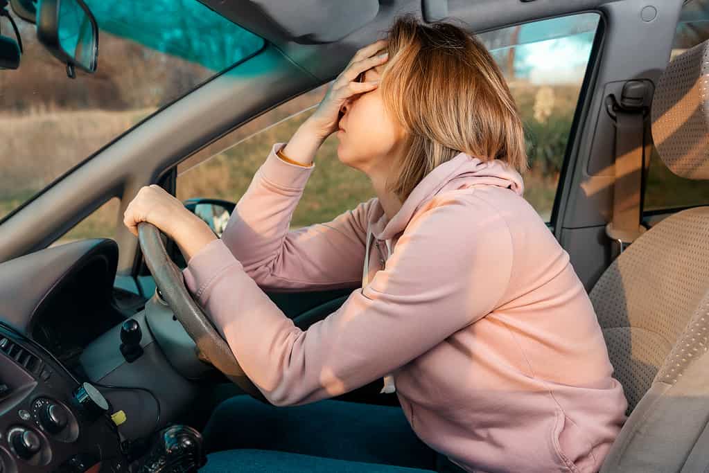 Una giovane donna bionda e triste è seduta al volante di un'auto e si tiene la testa disperata, coprendosi gli occhi con la mano.  Vista laterale.  Stress durante la guida di un'auto con guida a destra.