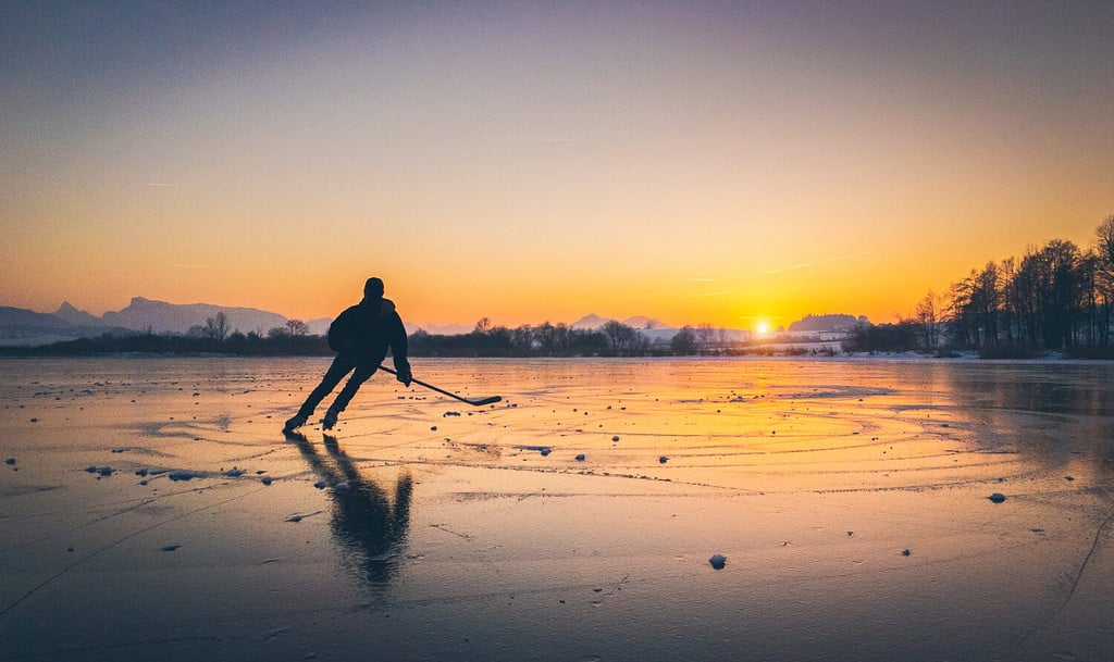 Vista panoramica panoramica della sagoma di un giovane giocatore di hockey che pattina su un lago ghiacciato con riflessi sorprendenti nella splendida luce dorata della sera al tramonto in inverno