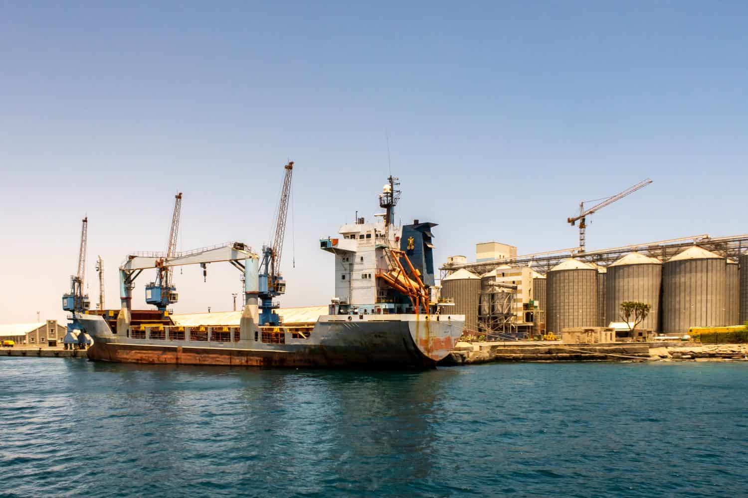 Grande nave portacontainer caricata con merci nel porto di Port Sudan.  Enormi gru portuali e fila di silos per il grano sullo sfondo.