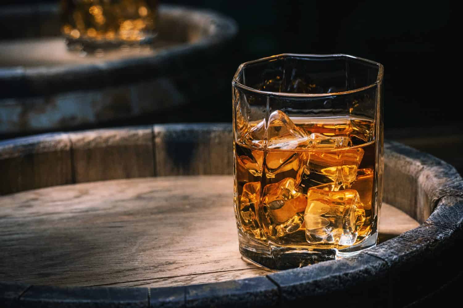Bottiglia e bicchiere di whisky con ghiaccio su un fondo di legno.  Un bicchiere di whisky scozzese e ghiaccio si trova sopra un barile di whisky rustico.  Whisky con ghiaccio.