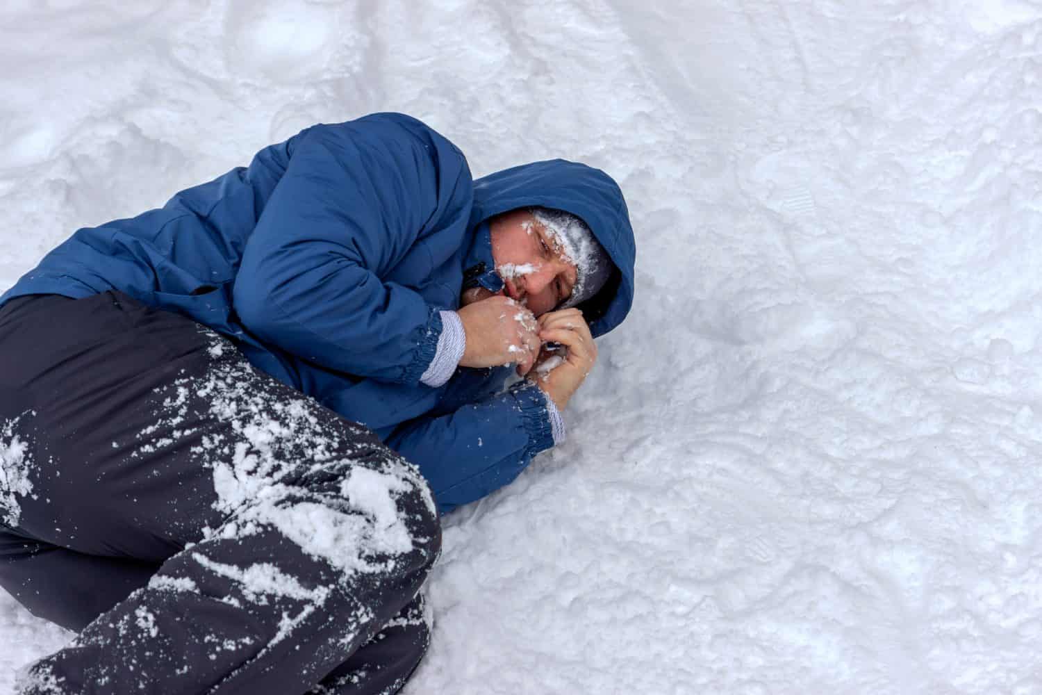 Un uomo congelato con una giacca blu e un cappello sdraiato coperto di neve e gelo, cercando di stare al caldo in una giornata invernale molto fredda, la neve cade intorno a lui.  Alpinista malato con ipotermia sulla neve durante il giorno.