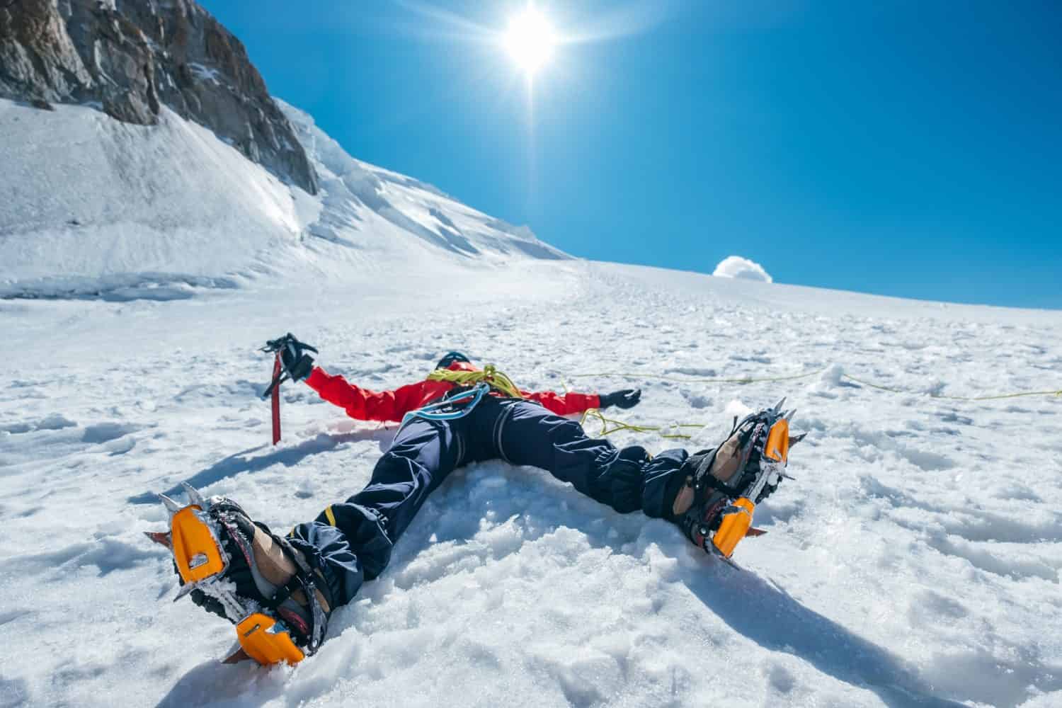 Scalatore esausto stanco che giace sotto il monte Mont Blanc du Tacul.  Gambe spalancate in stivali con ramponi e abiti da alpinismo con cielo azzurro con sfondo sole splendente.  Concetto di persone attive.