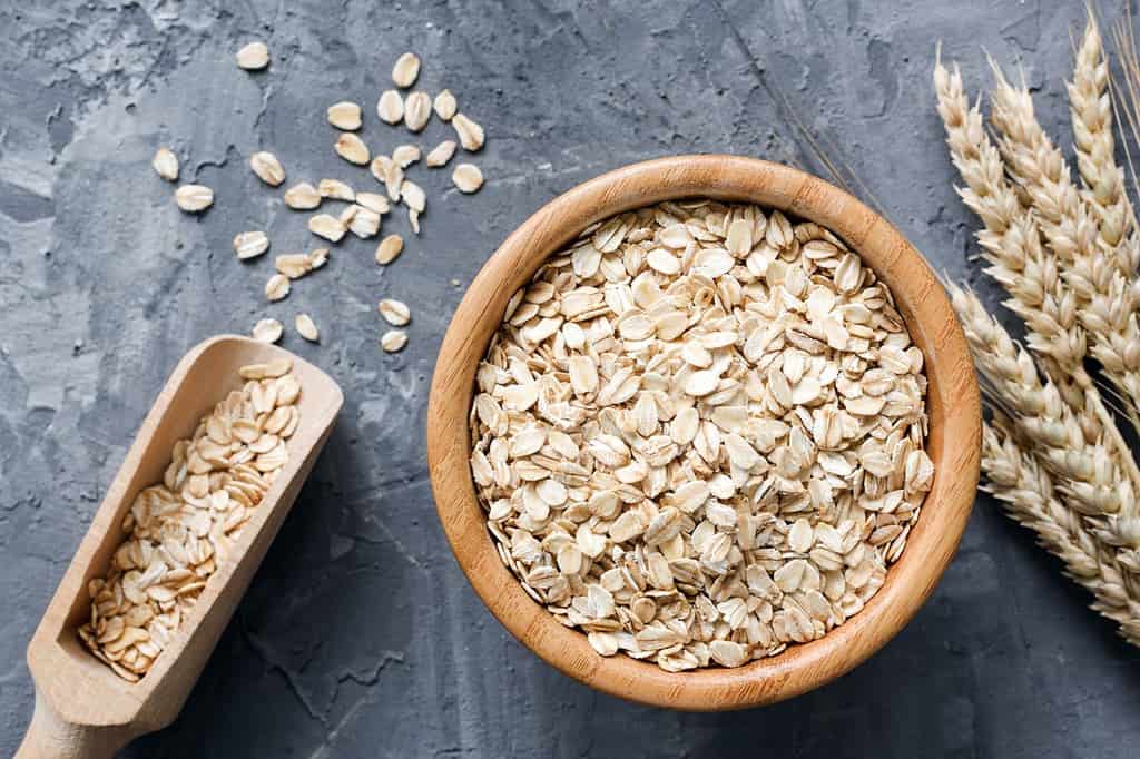 Fiocchi d'avena o fiocchi d'avena in ciotola di legno e spighe di grano dorate su fondo di pietra.  Vista dall'alto, orizzontale.  Stile di vita sano, alimentazione sana, concetto di cibo vegano