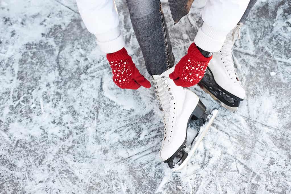 Ragazza che allaccia i lacci delle scarpe sui pattini da ghiaccio prima di pattinare sulla pista di pattinaggio, mani con guanti a maglia rossi.