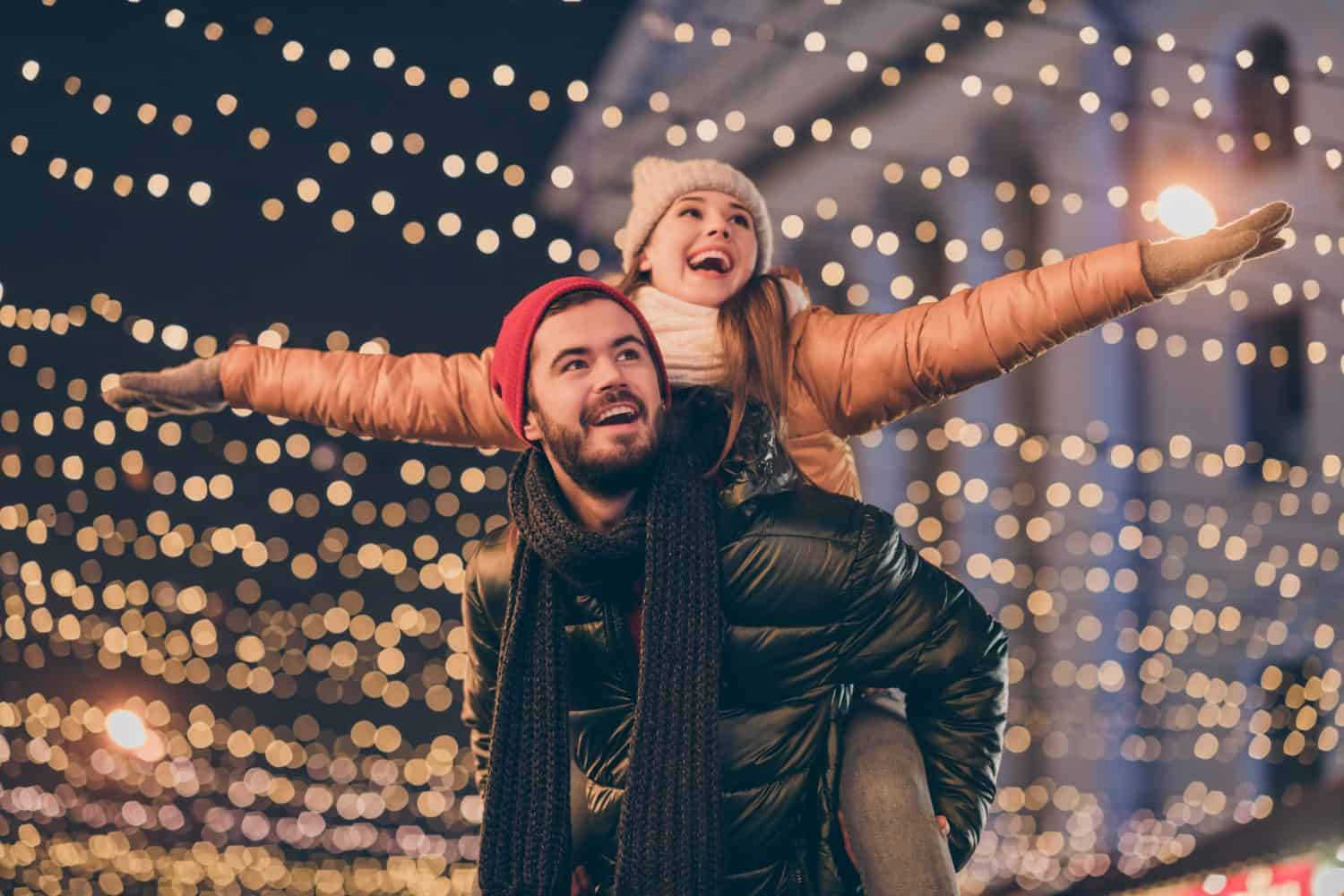 Foto di una coppia positiva che si diverte a Natale intorno alla sera all'esterno con illuminazione, il ragazzo sulle spalle e la ragazza che si tiene per mano volano