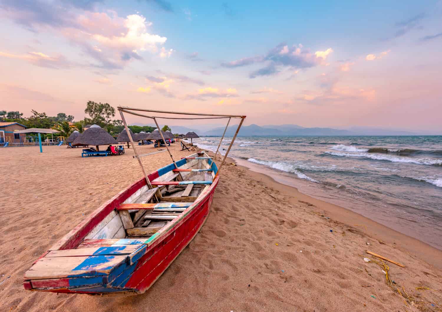 Burundi Bujumbura lago Tanganica, cielo nuvoloso ventoso e spiaggia di sabbia sul lago marino nell'Africa orientale, tramonto del Burundi con barca di legno.  Tetti africani di paglia sugli ombrelloni