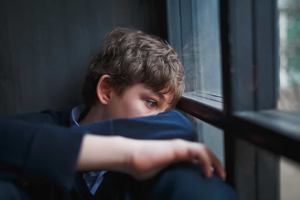 Adolescente pensieroso e triste con una camicia blu e jeans seduto alla finestra e chiude il viso con le mani.