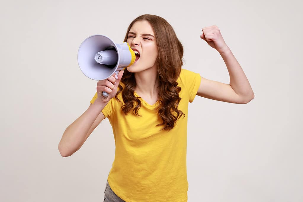 Una ragazza adolescente arrabbiata e nervosa con i capelli castani in maglietta gialla che urla ad alta voce al megafono, fa annunci, protesta, vuole essere ascoltata.