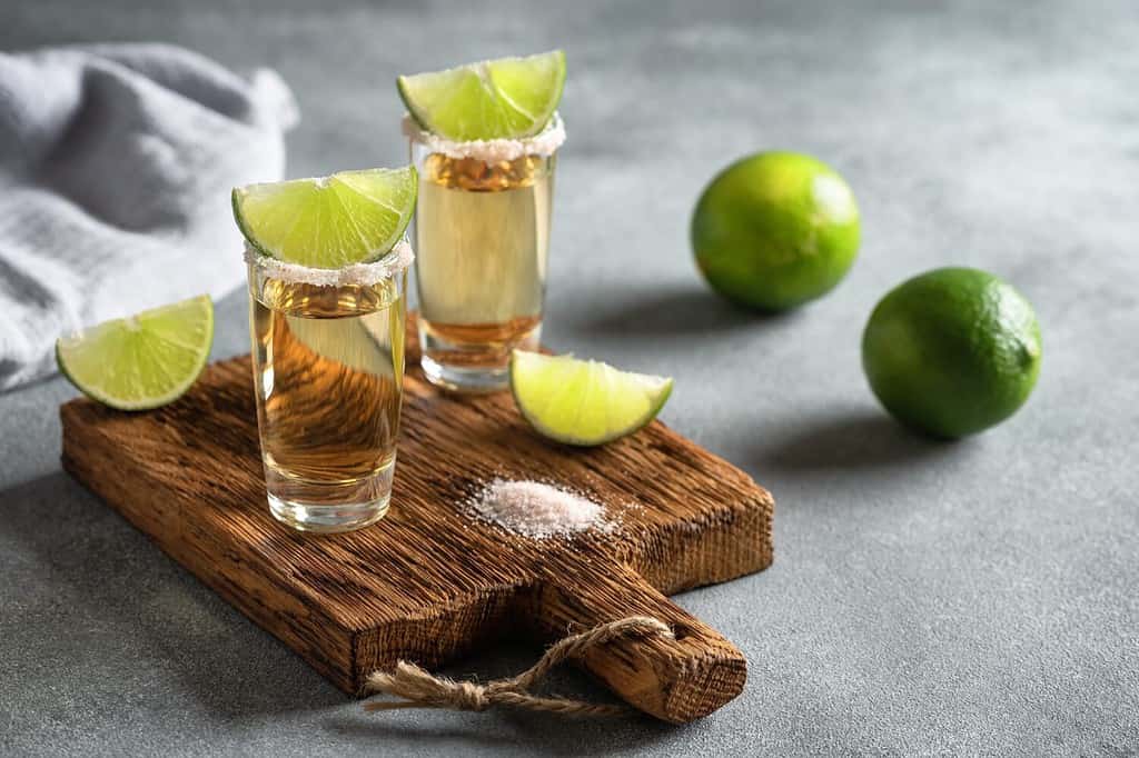 Tequila messicana d'oro in un bicchierino con lime e sale rosa su una tavola di legno, sfondo grigio cemento.  Vista laterale, messa a fuoco selettiva.