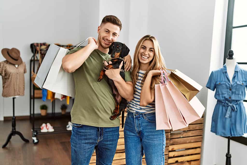 Giovane coppia di clienti ispanici che sorride felice tenendo le borse della spesa e il cane al negozio di abbigliamento.
