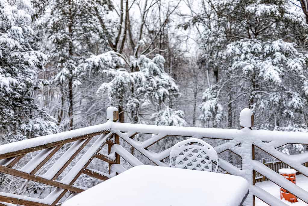 Sedia da tavolo per esterni in metallo bianca coperta di neve sul ponte di legno della casa con recinzione ringhiera in inverno con alberi forestali nel nord della Virginia