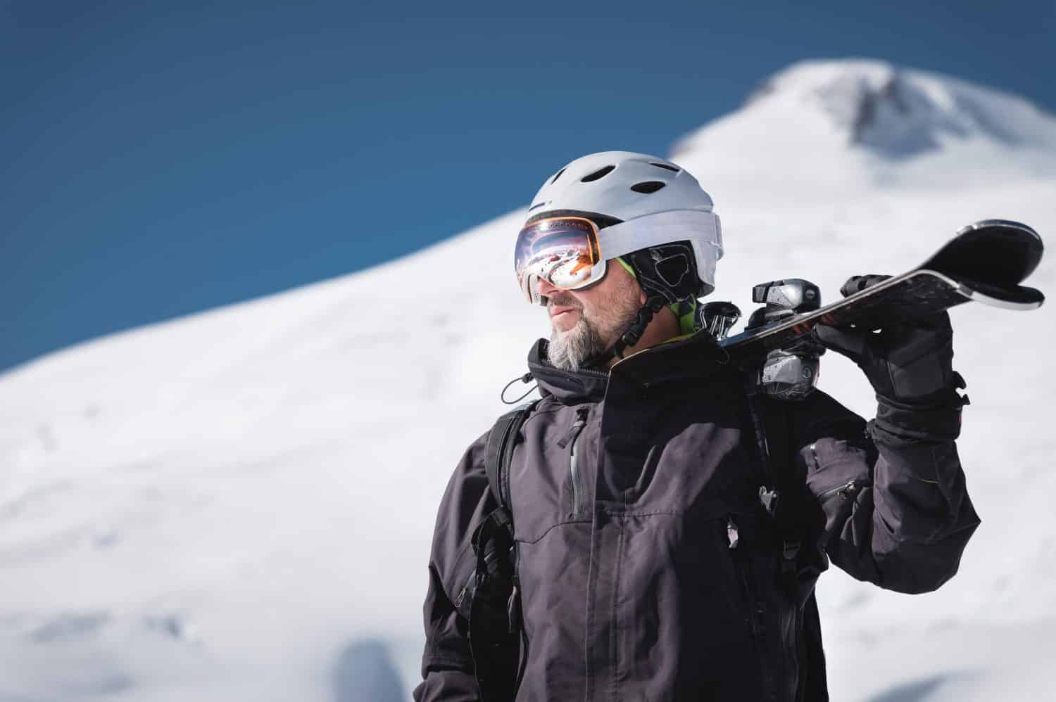 Ritratto barbuto sciatore maschio invecchiato sullo sfondo delle montagne innevate del Caucaso.  Un uomo adulto che indossa una maschera da sci e un casco da sci sulla spalla guarda le montagne.  Concetto di stazione sciistica