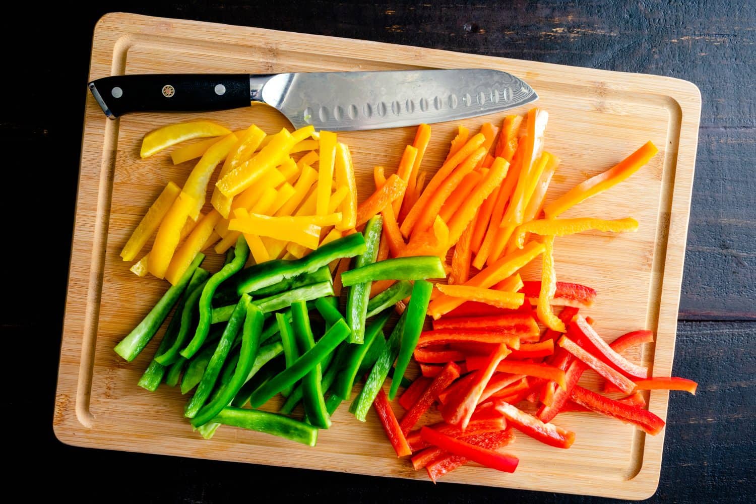 Peperoni rossi, arancioni, gialli e verdi tagliati a strisce sottili: peperoni tagliati a batonnet su un tagliere di bambù con un coltello da chef