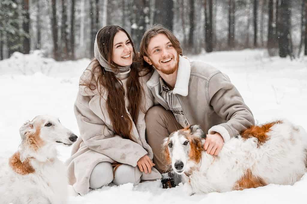 Adoro una giovane coppia romantica, un ragazzo nella foresta invernale fredda e nevosa che cammina con un animale domestico, un cane di razza borzoi russo da caccia.  Levriero, proprietario di levrieri.  Divertirsi, ridere.  Pelliccia elegante, cappello di lana.