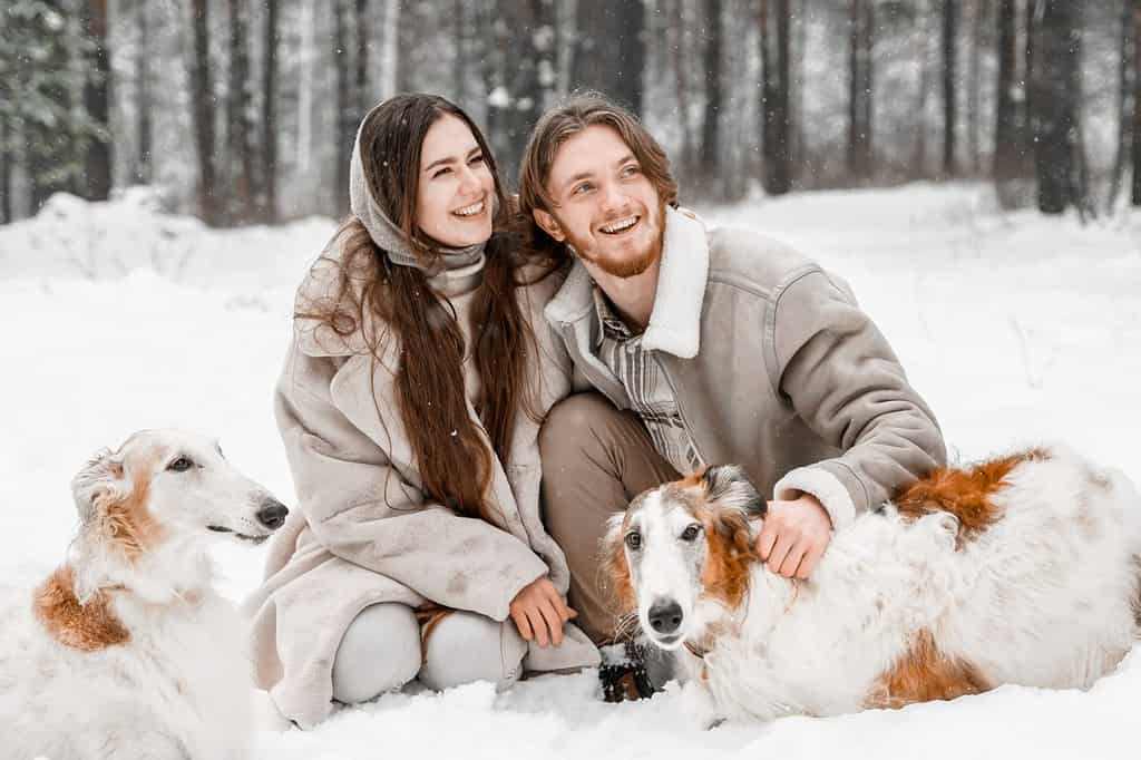 Adoro una giovane coppia romantica, un ragazzo nella foresta invernale fredda e nevosa che cammina con un animale domestico, un cane di razza borzoi russo da caccia.  Levriero, proprietario di levrieri.