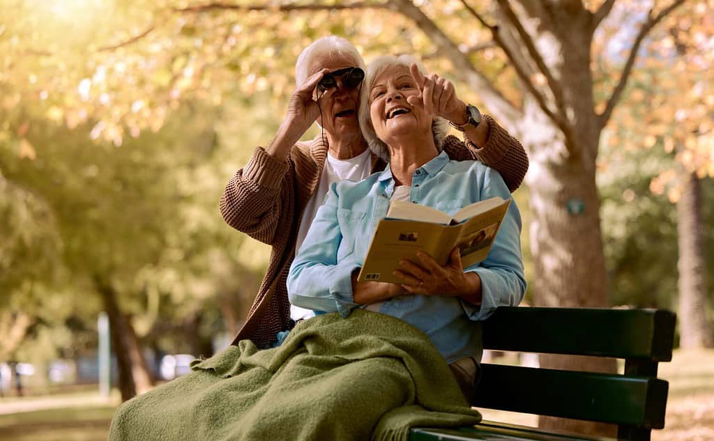 Parco, panchina e binocolo con una coppia senior che osserva gli uccelli insieme all'aperto nella natura durante l'estate.  Primavera, amore e libro con un uomo maturo e una donna che legano mentre sono seduti in un giardino per ammirare il panorama