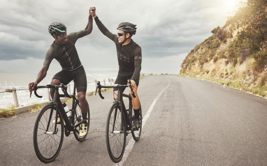 Bici, amici e uomini danno il cinque su strada divertendosi a pedalare insieme all'aperto.  Successo, diversità e lavoro di squadra dei ciclisti maschi in bicicletta su strada, esercizio fisico o allenamento su asfalto.