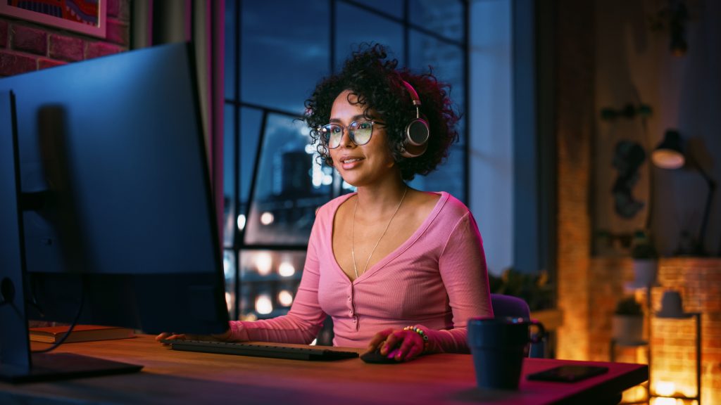 Giovane bella donna nera in cuffie che utilizza il computer in un elegante loft la sera.  Sorridere femminile creativo, sfogliare video sui social media.  Vista urbana della città dalla grande finestra.