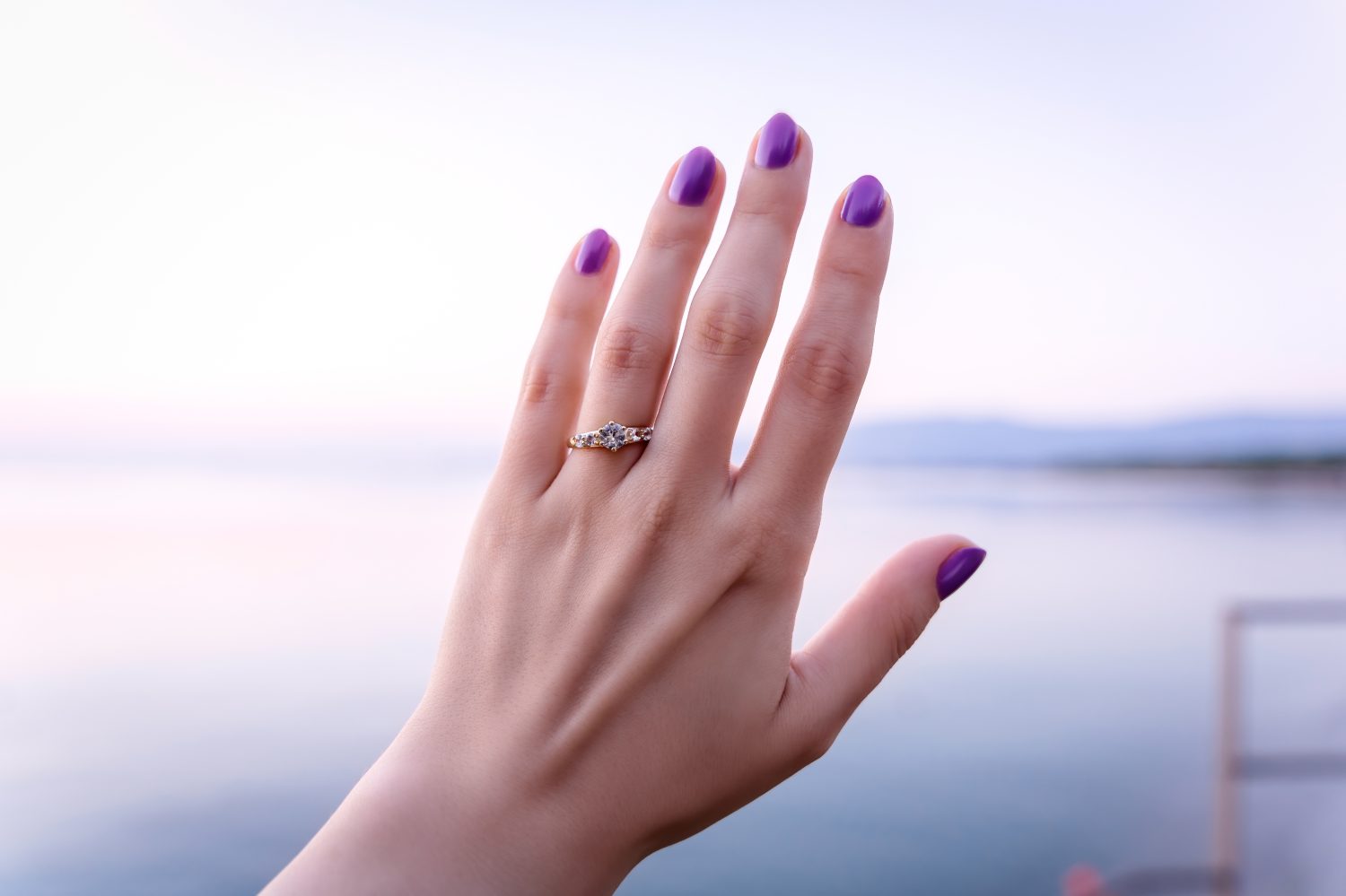 Mano di donna con una proposta di anello con diamanti in riva al mare, proposta di alba sulla spiaggia, anello dorato con zaffiro bianco