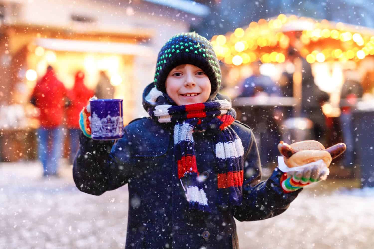 Un ragazzino carino che mangia salsiccia tedesca e beve un pugno caldo per bambini sul mercatino di Natale.  Bambino felice sul tradizionale mercato familiare in Germania.  Ragazzo che ride in abiti invernali colorati