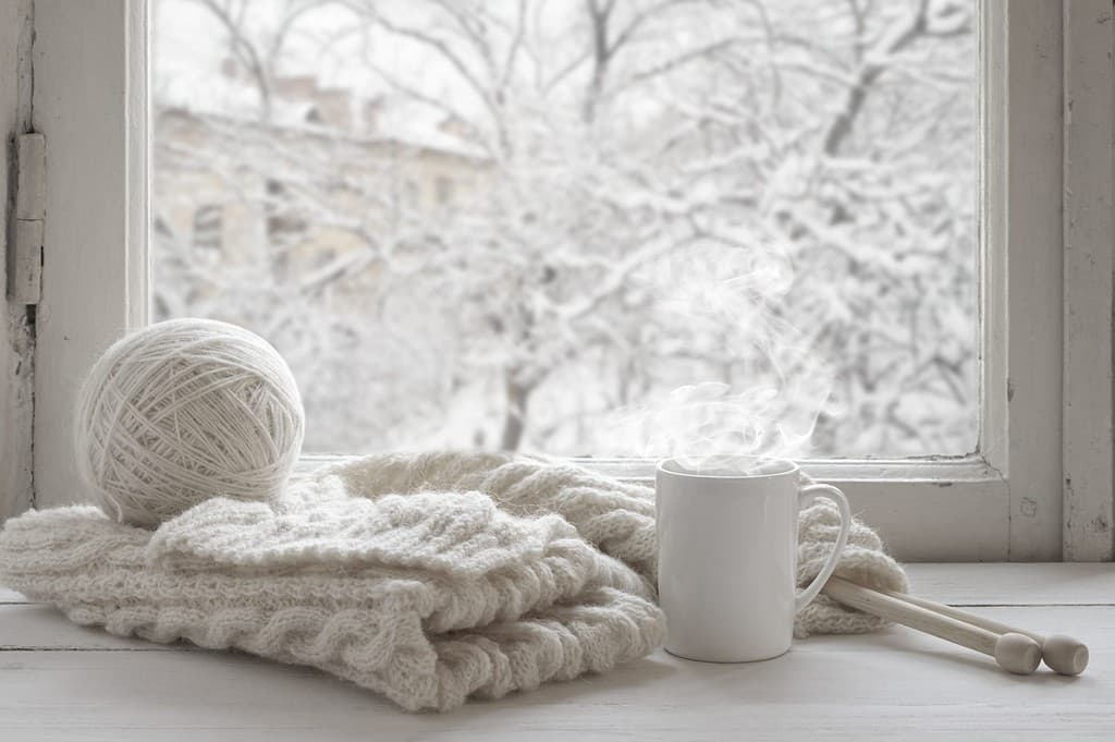 Accogliente natura morta invernale: tazza di tè caldo e calda maglia di lana sul davanzale vintage contro il paesaggio innevato dall'esterno.