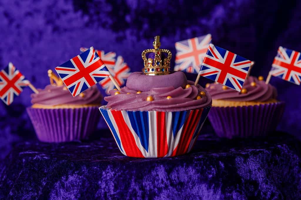Cupcakes per l'incoronazione reale per celebrare l'incoronazione di re Carlo III.  Cupcake decorati con la corona, sfondo di velluto viola, bandiere dell'Union Jack, cupcake di lusso su un piedistallo.