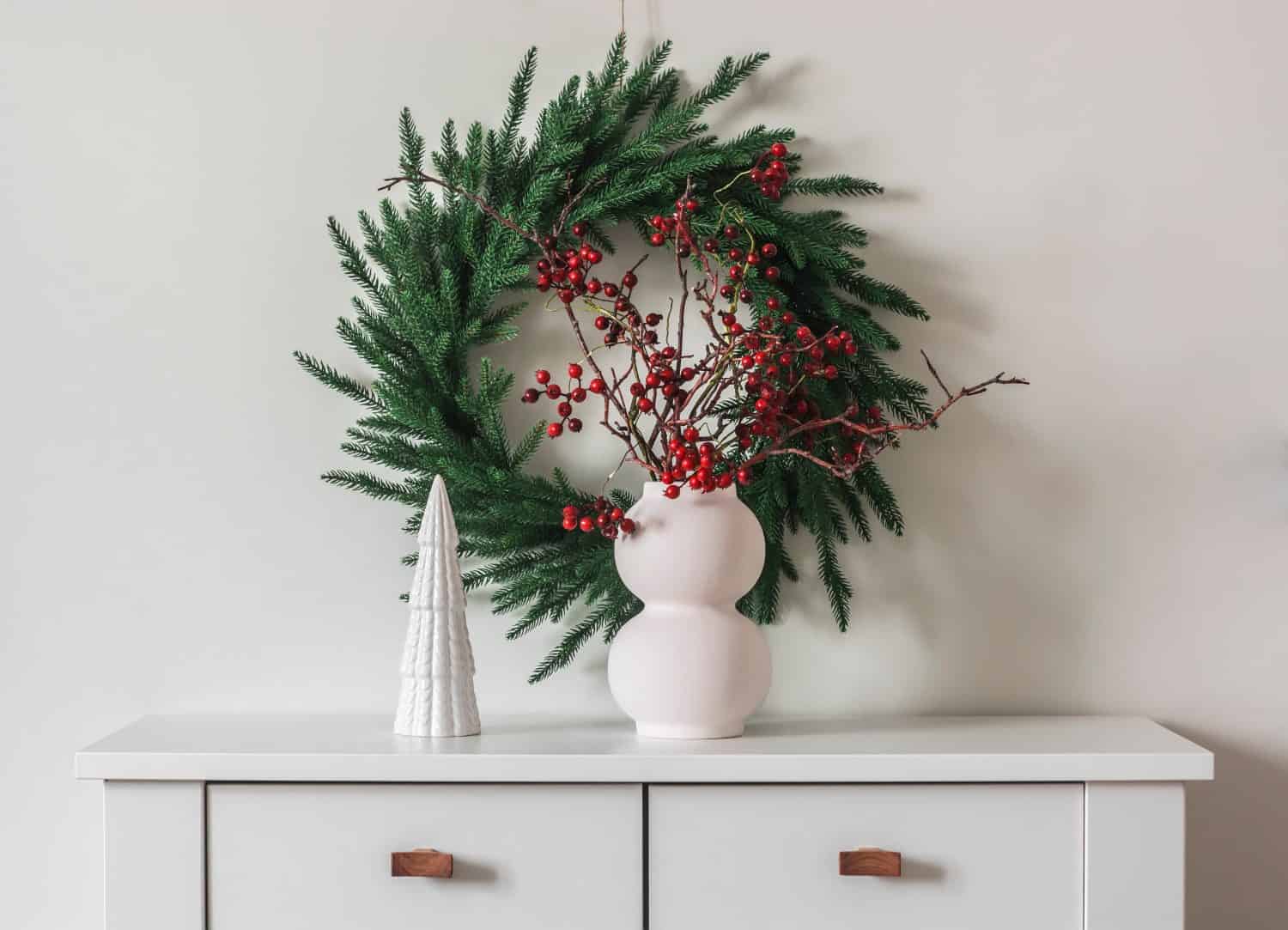 Decorazioni natalizie in stile scandinavo nel soggiorno: una ghirlanda di abete rosso sul muro, un vaso con rami di mirtillo rosso su una cassettiera bianca