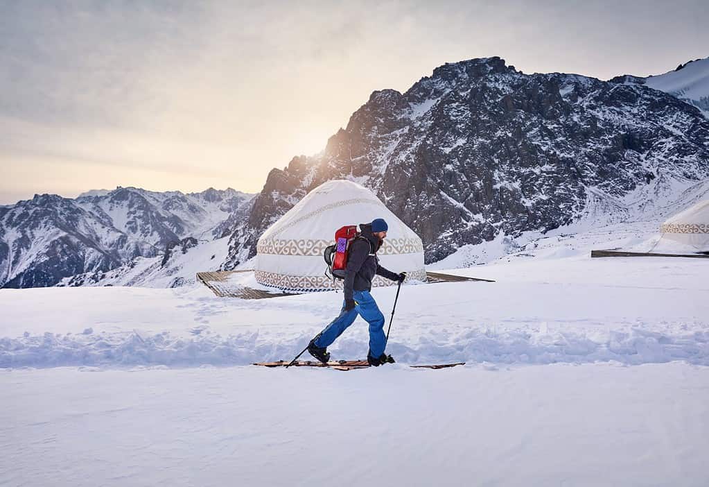 Sciatore uomo sci alpinismo in salita con barba e zaino in alta montagna innevata vicino alla tradizionale casa yurta nomade nella stazione sciistica Shymbulak ad Almaty, Kazakistan.  Sport all'aria aperta in backcountry stagione invernale.