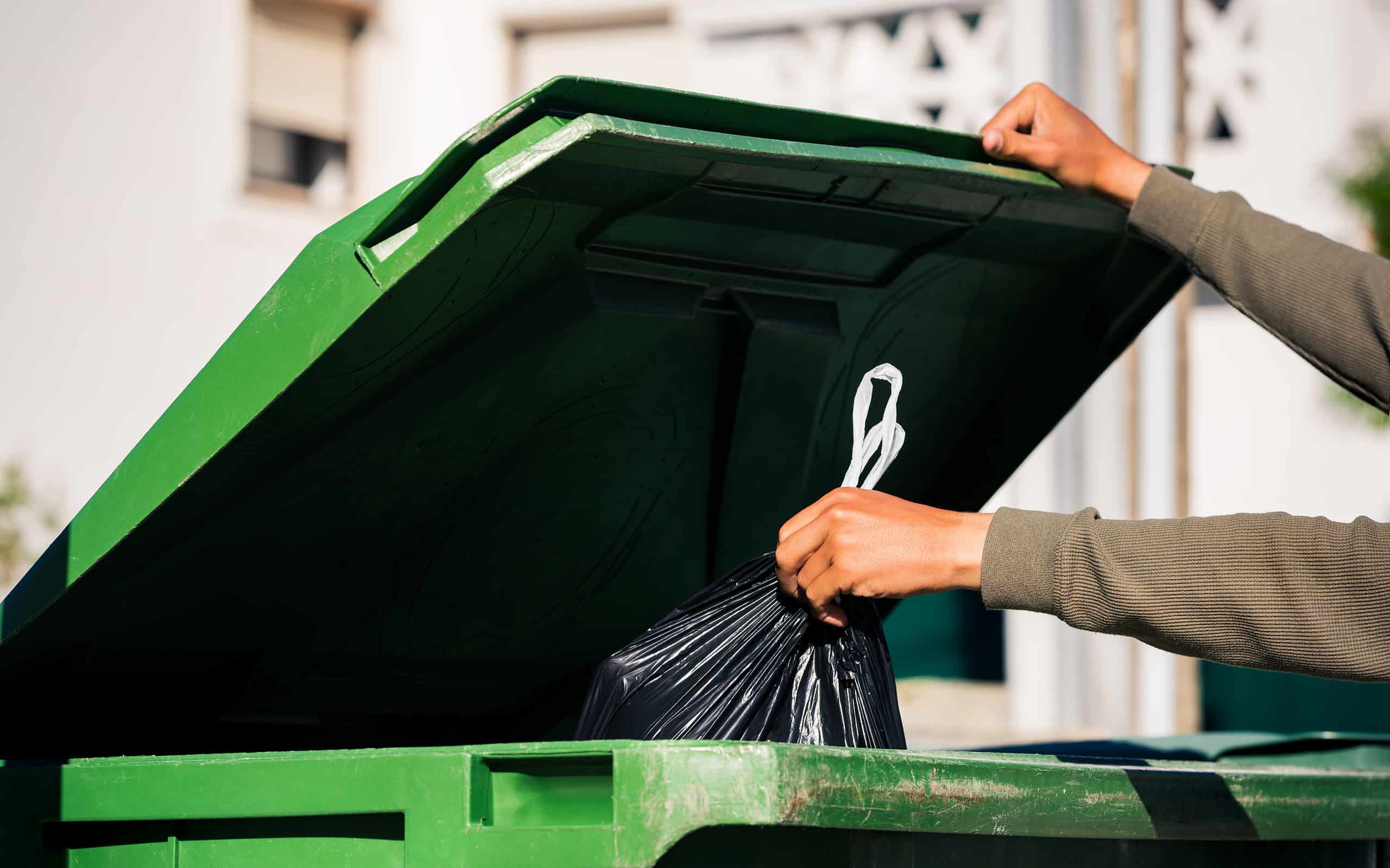 Uomo che butta fuori il sacchetto della spazzatura nero ecologico riciclabile in un grande contenitore per rifiuti verde in plastica.  Porta fuori la spazzatura