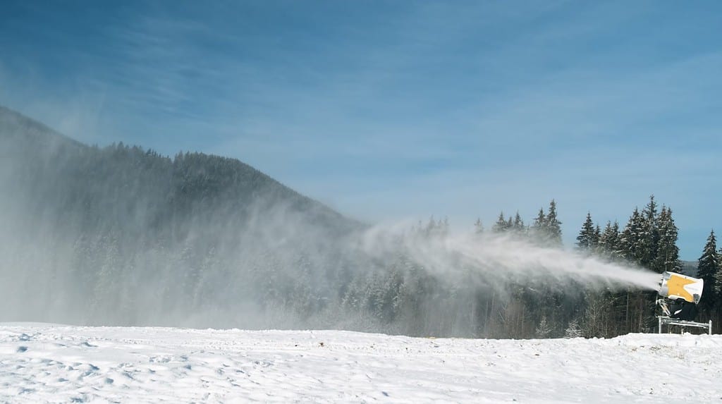 Il cannone da neve produce neve artificiale in una luminosa giornata di sole nella stazione sciistica.  Sul retro foresta invernale, cielo blu.  Gli impianti di innevamento spruzzano acqua per produrre neve.  Preparazione della pista da sci per lo sci, gli sport invernali
