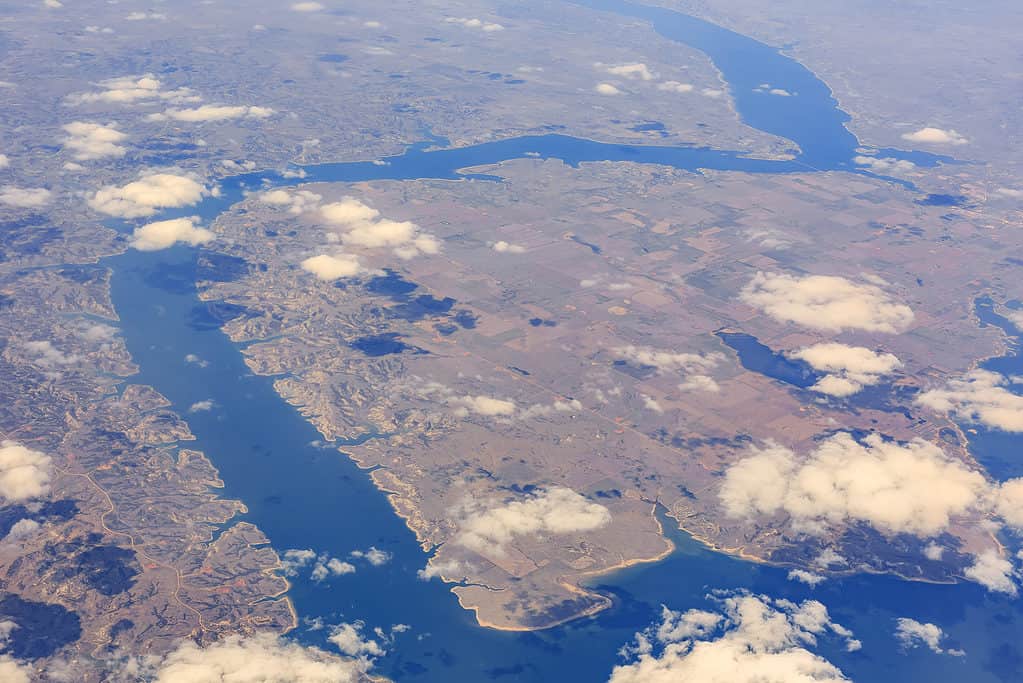 Una veduta aerea del lago Sakakawea.  Il lago è stretto e serpeggia attraverso l'inquadratura dall'inquadratura in basso a destra attraverso la parte sinistra della fotografia e poi torna a destra per circa 3/4 della salita e poi in alto verso destra ci sono alcune nuvole tra la macchina fotografica e la fotografia e il treno è troppo lontano per sembrare altro che marrone e verde.