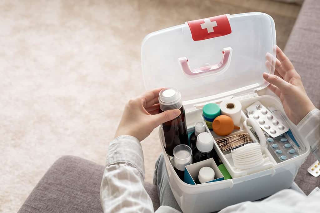 Primo piano della mano femminile che posiziona ordinatamente il medicinale nella vista dall'alto del kit di pronto soccorso domestico.  Organizzazione di stoccaggio in scatola di plastica trasparente per farmaci, pillole, siringhe, bende.  Fornitura rapida di emergenza per la sicurezza dell'aiuto sanitario