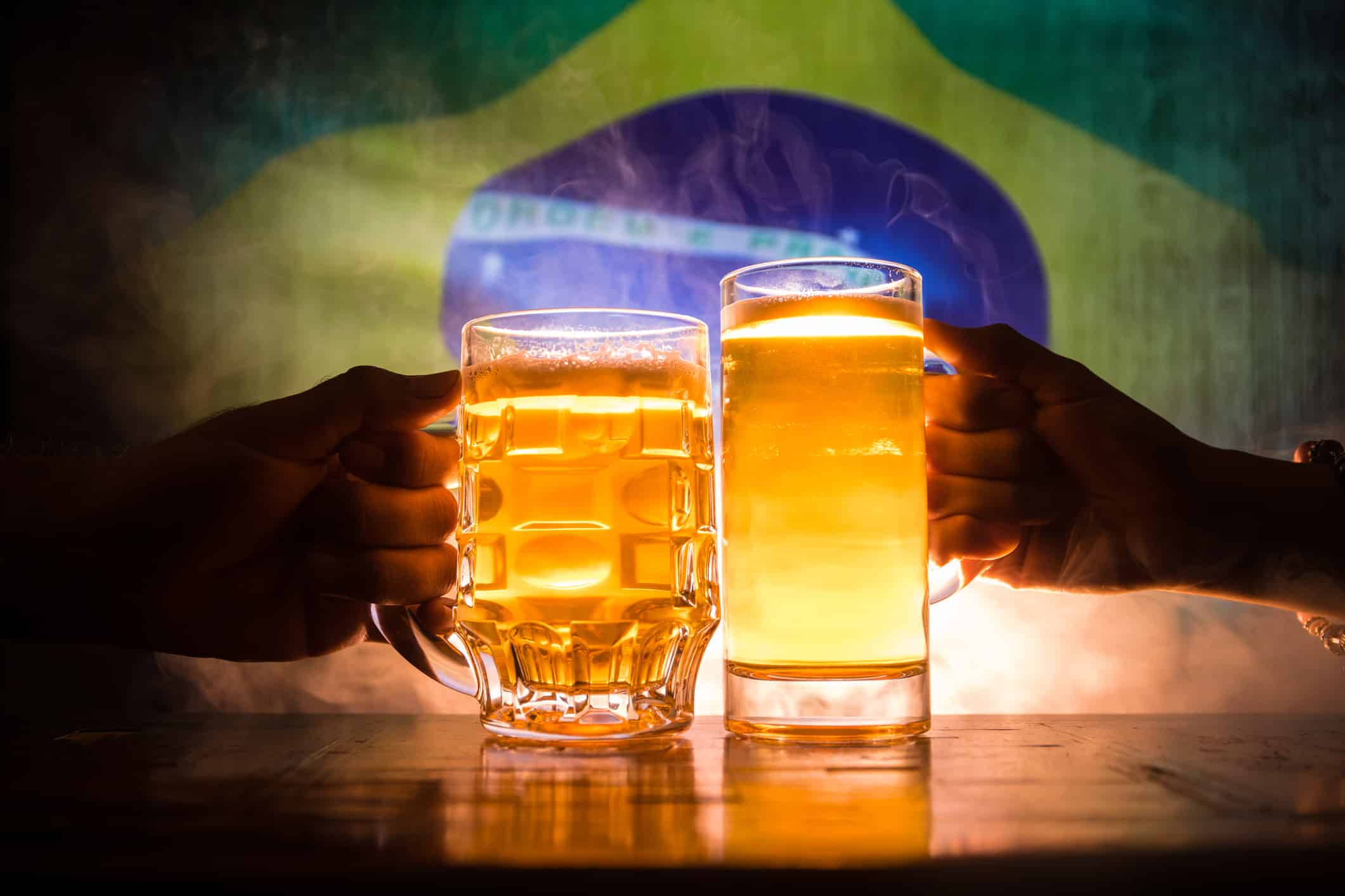 Due amici brindano (tintinnano) con bicchieri di birra leggera al pub.  Bellissimo sfondo con vista offuscata della bandiera del Brasile.  Sostieni il tuo paese con il concetto di birra.