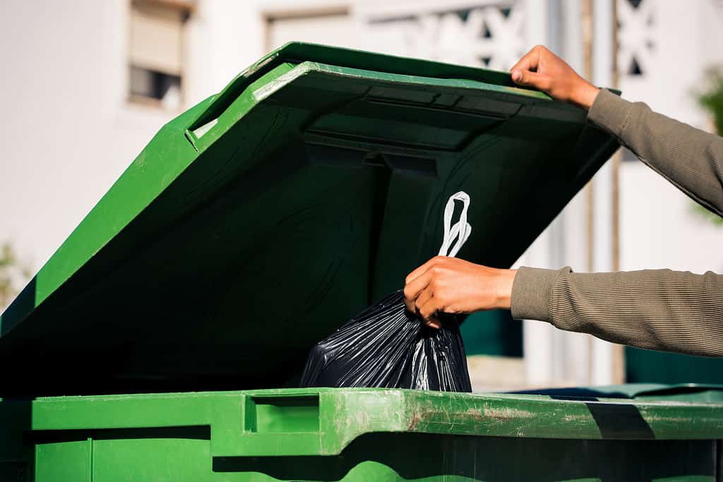 Uomo che butta fuori il sacchetto della spazzatura nero ecologico riciclabile in un grande contenitore per rifiuti verde in plastica.  Porta fuori la spazzatura