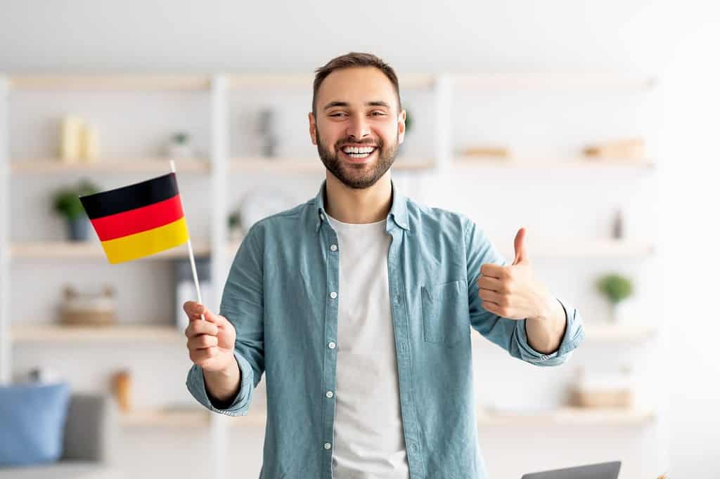 Felice uomo caucasico che mostra il pollice in su e la bandiera della Germania, in posa e sorridente alla telecamera in ambienti chiusi.  Studente millenario allegro che consiglia l'istruzione straniera, imparando la lingua tedesca
