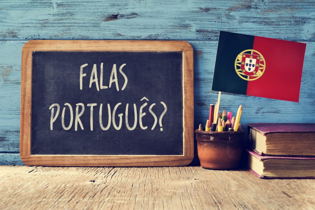 una lavagna con la domanda question falas portuges?  parli portoghese?  scritta in portoghese, un vaso con matite, alcuni libri e la bandiera del Portogallo, su una scrivania di legno