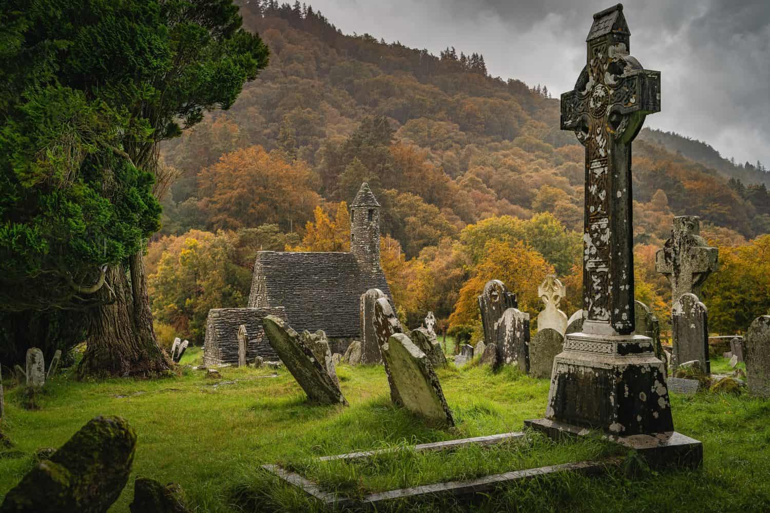 Tombe antiche della chiesa medievale Croci celtiche nel cimitero di Glendalough.  Foresta autunnale lunatica, montagne sotto la pioggia, cielo tempestoso sullo sfondo Wicklow Irlanda