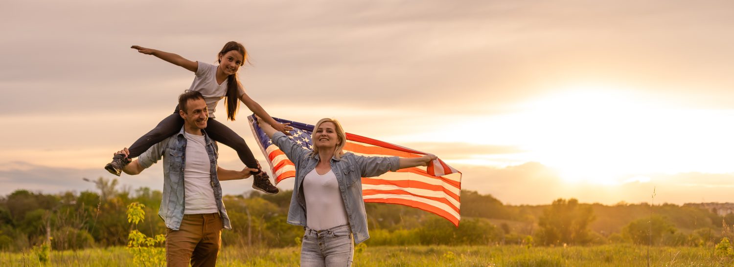 Giovani genitori con la figlia che tiene la bandiera americana in campagna al tramonto.  Celebrazione del Giorno dell'Indipendenza
