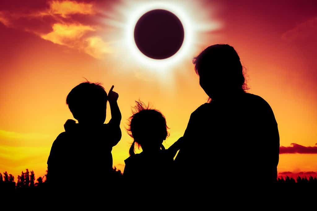 Silhouette vista posteriore della famiglia seduta e rilassante insieme.  Il ragazzo indica l'eclissi solare sullo sfondo del cielo dorato.  Famiglia felice che trascorre del tempo insieme.  All'aperto.