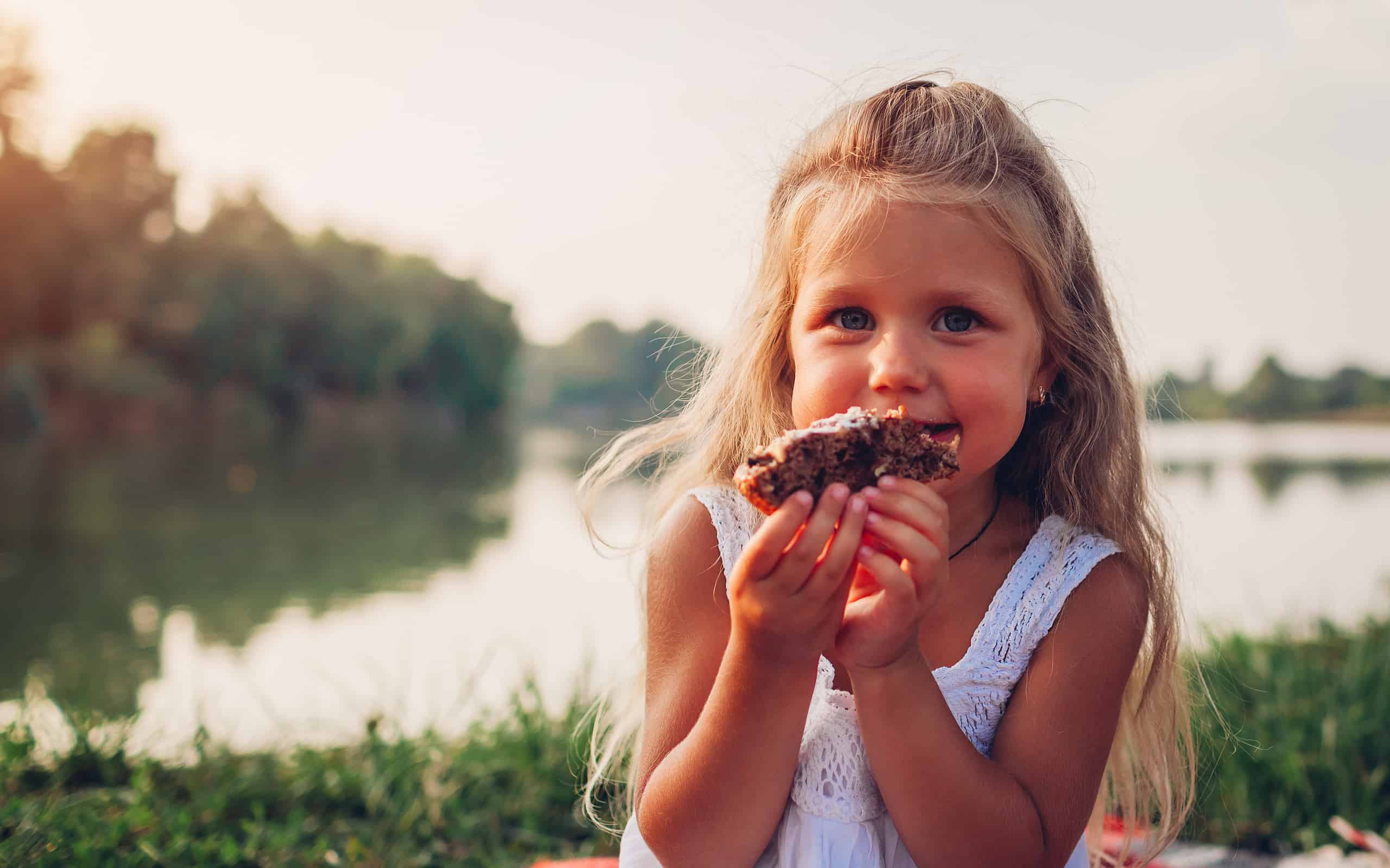Bambina che mangia torta durante un picnic in famiglia vicino al fiume estivo.  Bambino che tiene pezzo di torta