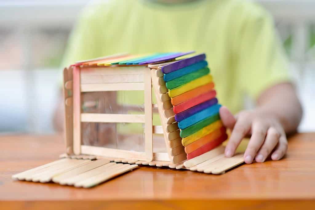 Bambino asiatico con la casa dei bastoncini di ghiacciolo all'aperto. Giovane maschio con innovazione creativa in legno. Bambino che fa un mestiere di design modello colorato. Progetto di attività fatta a mano per ragazzo homeschooling.